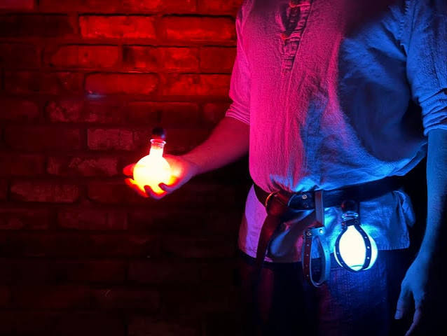 A person in larp costume holds a potion bottle shaped red light. Another potion bottle shaped light hangs in a leather holster on their belt.