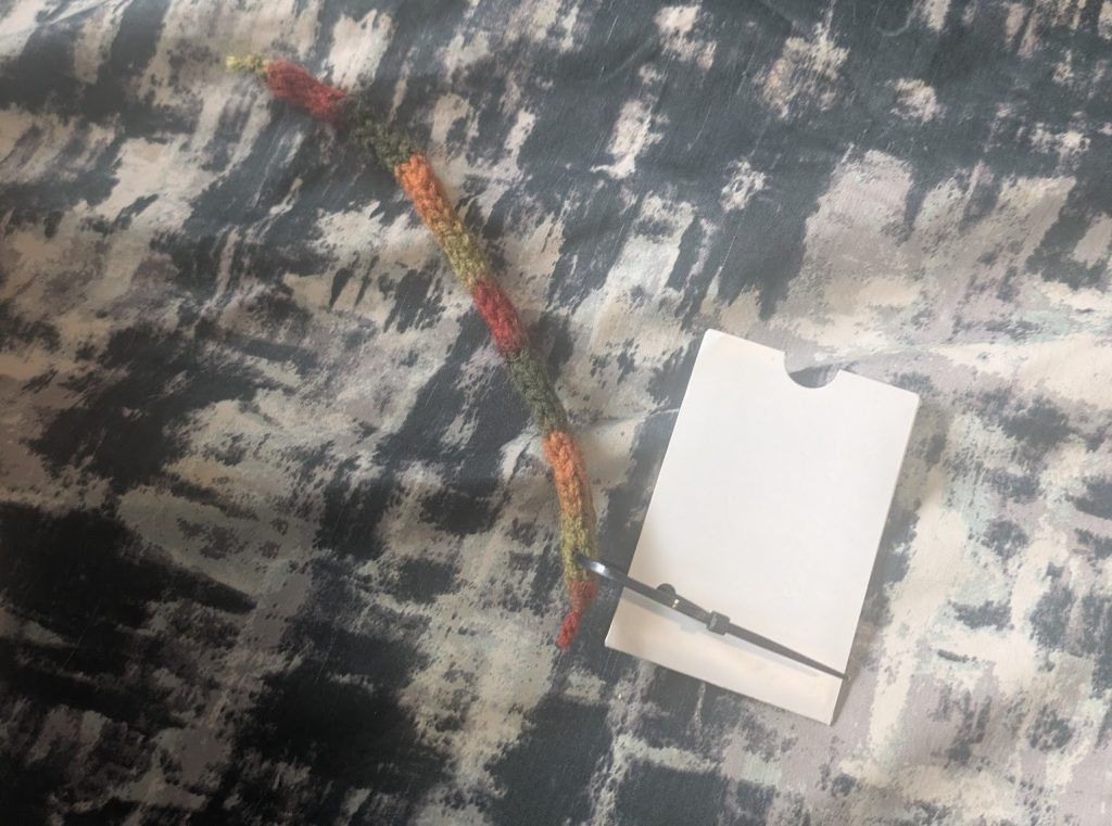 A length of i-cord in stripes of red green and orange wool attached to a lammie with a cable tie near one end of the i-cord. It is approximately one and a half times the length of the lammie