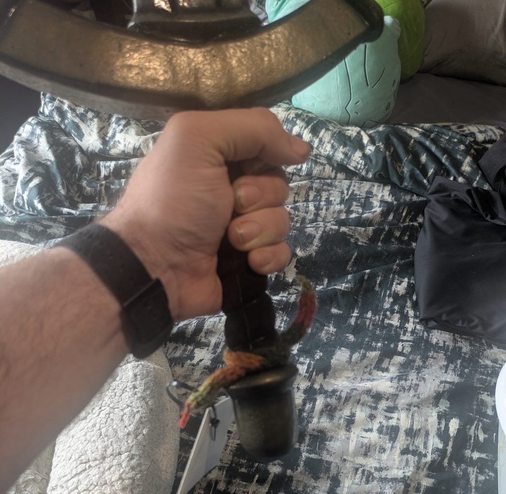 The striped woolen i-cord from the previous image is tied around the hilt a larp sword. The sword is held aloft showing the lammie hanging beside the bottom of the hilt